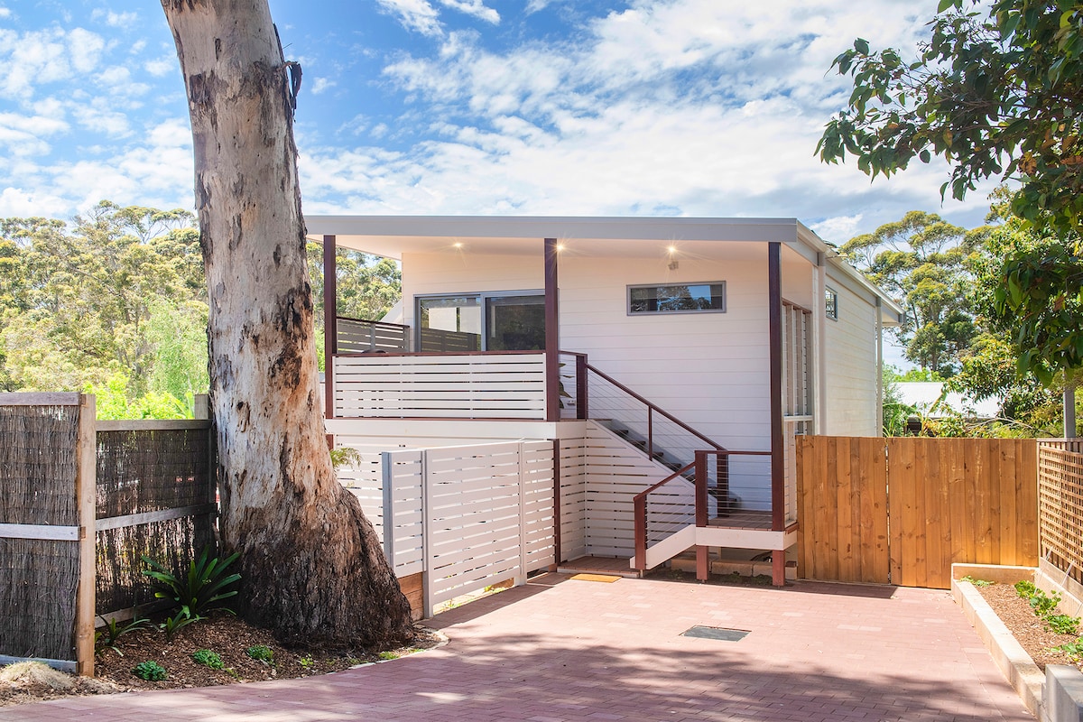 A private studio apartment is situated at the base of a tall Karri tree, with a staircase leading up to the entrance. The exterior showcases a clean and modern design, complemented by a wooden fence and paved driveway.