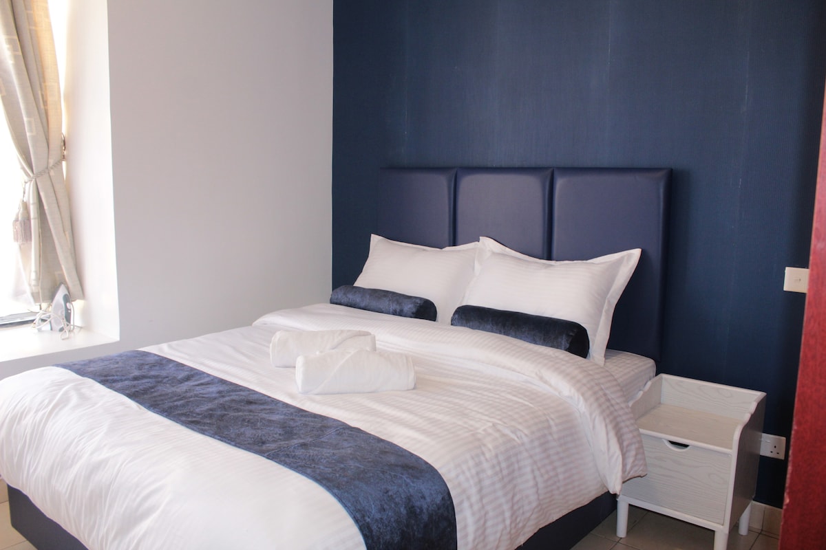 A spacious bed is prominently positioned, adorned with crisp white linens and a deep blue accent throw. Side tables with a minimalistic design complement the serene atmosphere. Natural light filters through a window, enhancing the calming tones of the interior.