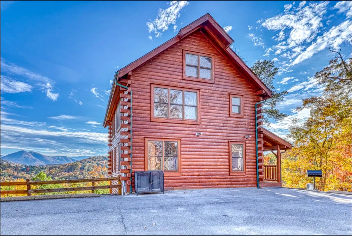 The two-story wooden cabin features a rustic design and multiple large windows, showcasing panoramic views of the surrounding mountains. The exterior is complemented by a spacious driveway and wooden railings, while vibrant autumn foliage frames the building, highlighting its natural setting.