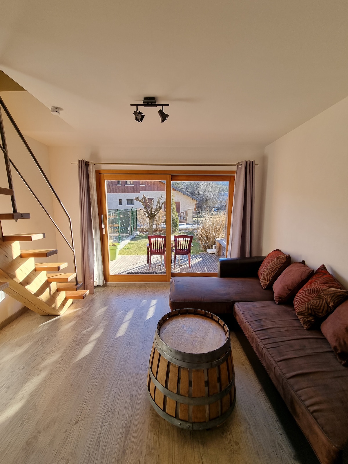A cozy living area is visible with a comfortable brown couch adorned with decorative pillows. A wooden barrel coffee table is placed in front. Large sliding glass doors provide a view of the garden, where two chairs are arranged on a wooden deck.