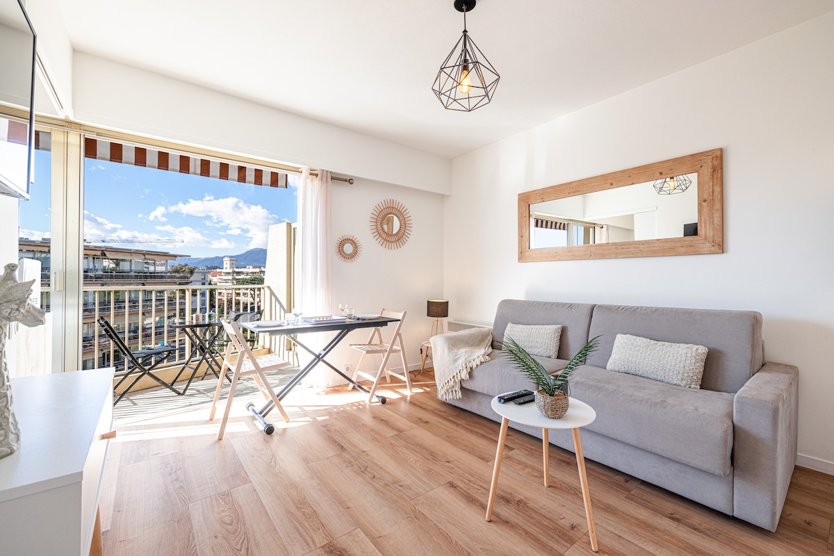 The living area is furnished with a comfortable sofa facing an open terrace. A small round table and folding chairs are positioned nearby, while a decorative mirror enhances the space. Natural light fills the room through large glass doors, leading to views of the outdoor balcony.