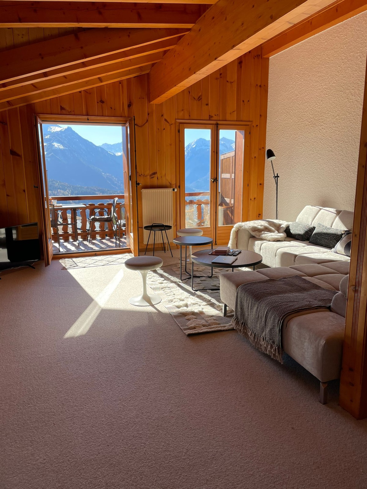 A spacious living area features wooden walls and a sloped ceiling, allowing natural light to fill the room. Large windows provide views of the surrounding mountains. A comfortable sofa is positioned near a coffee table, accompanied by modern chairs. A decorative lamp stands nearby.