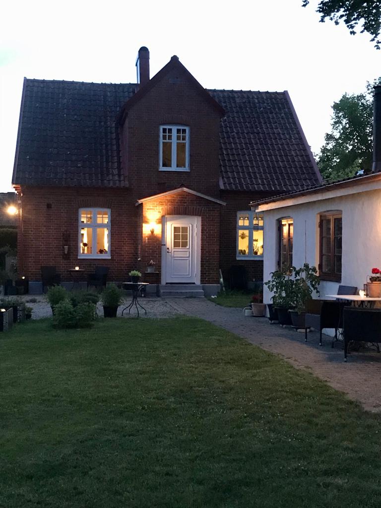 A charming two-story brick house is illuminated by soft outdoor lighting, showcasing a welcoming front door and decorative windows. Surrounding the home, a well-maintained lawn is complemented by patio furniture, creating inviting outdoor spaces for relaxation.