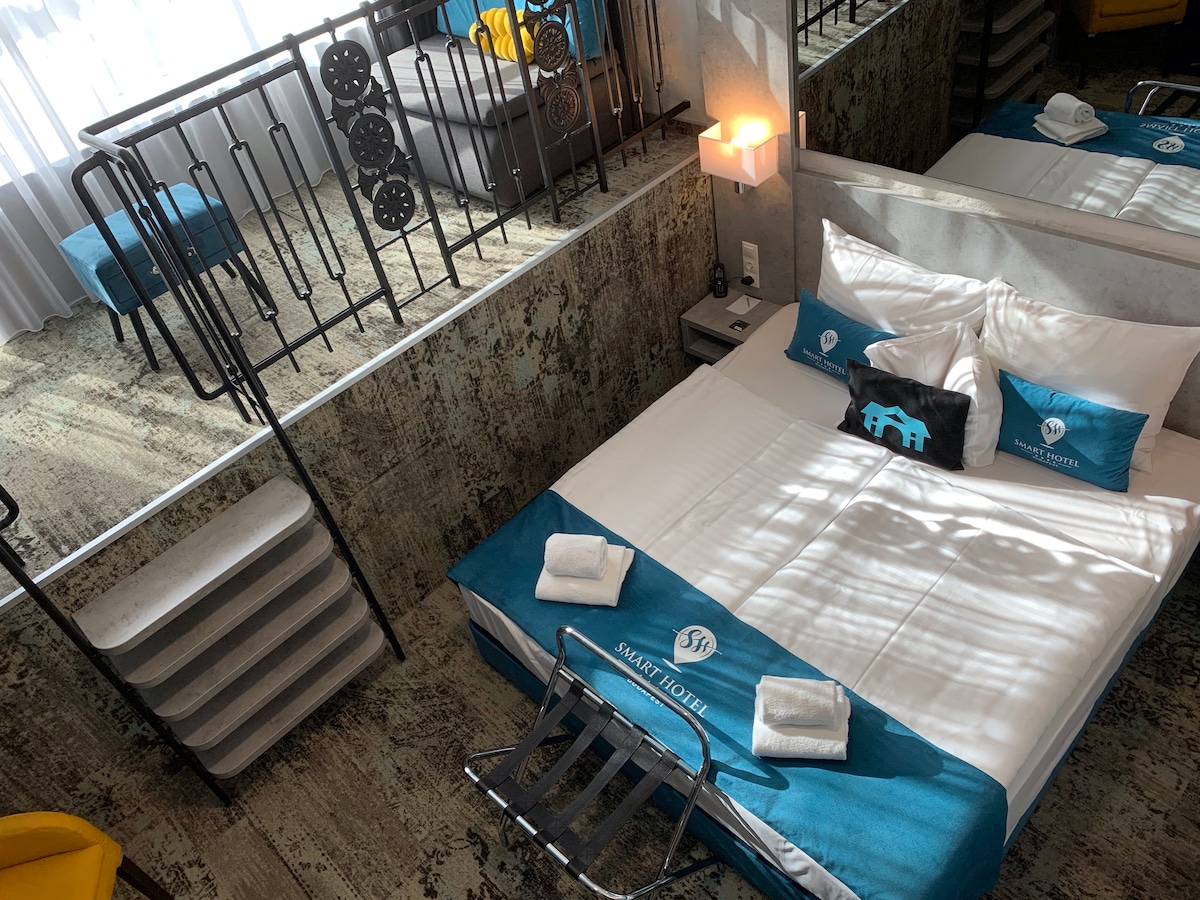 A spacious sleeping area is highlighted by a large bed dressed in crisp white linens and accented with turquoise pillows. A soft area rug adds warmth to the floor, and a modern seating area is visible in the background. Natural light floods the room through a large window.