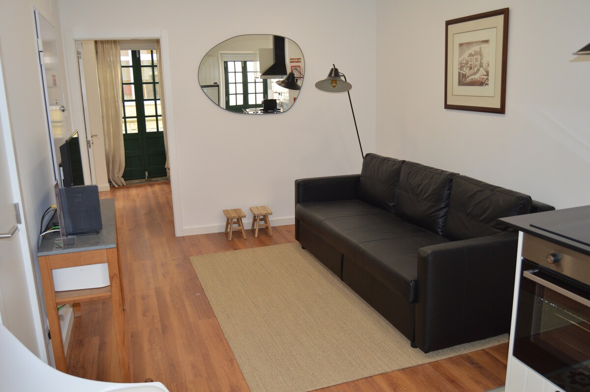 A living area is presented with a black leather sofa facing a flat-screen television. A round mirror is mounted on the wall, reflecting the space. A light-colored area rug is laid on the wooden floor, and two small stools are positioned nearby.