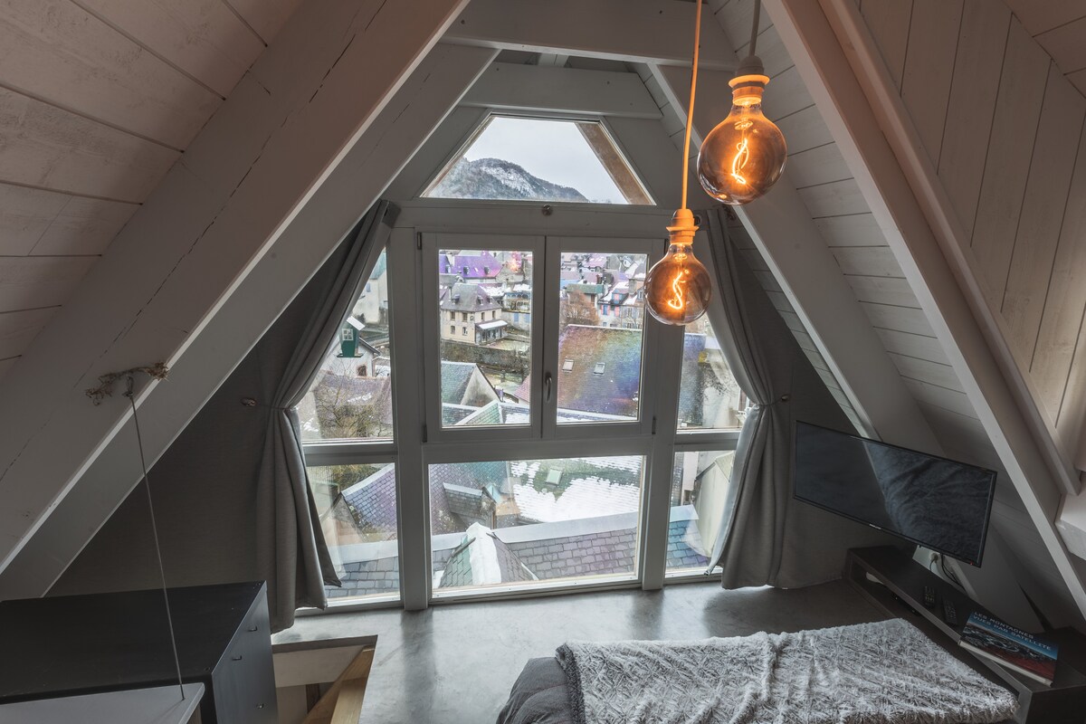 A loft-style bedroom features large triangular windows that offer expansive views of the surrounding village and mountains. Soft grey curtains frame the windows, and modern pendant lights hang from the sloped ceiling. A TV is positioned on a low unit beside a spacious bed.