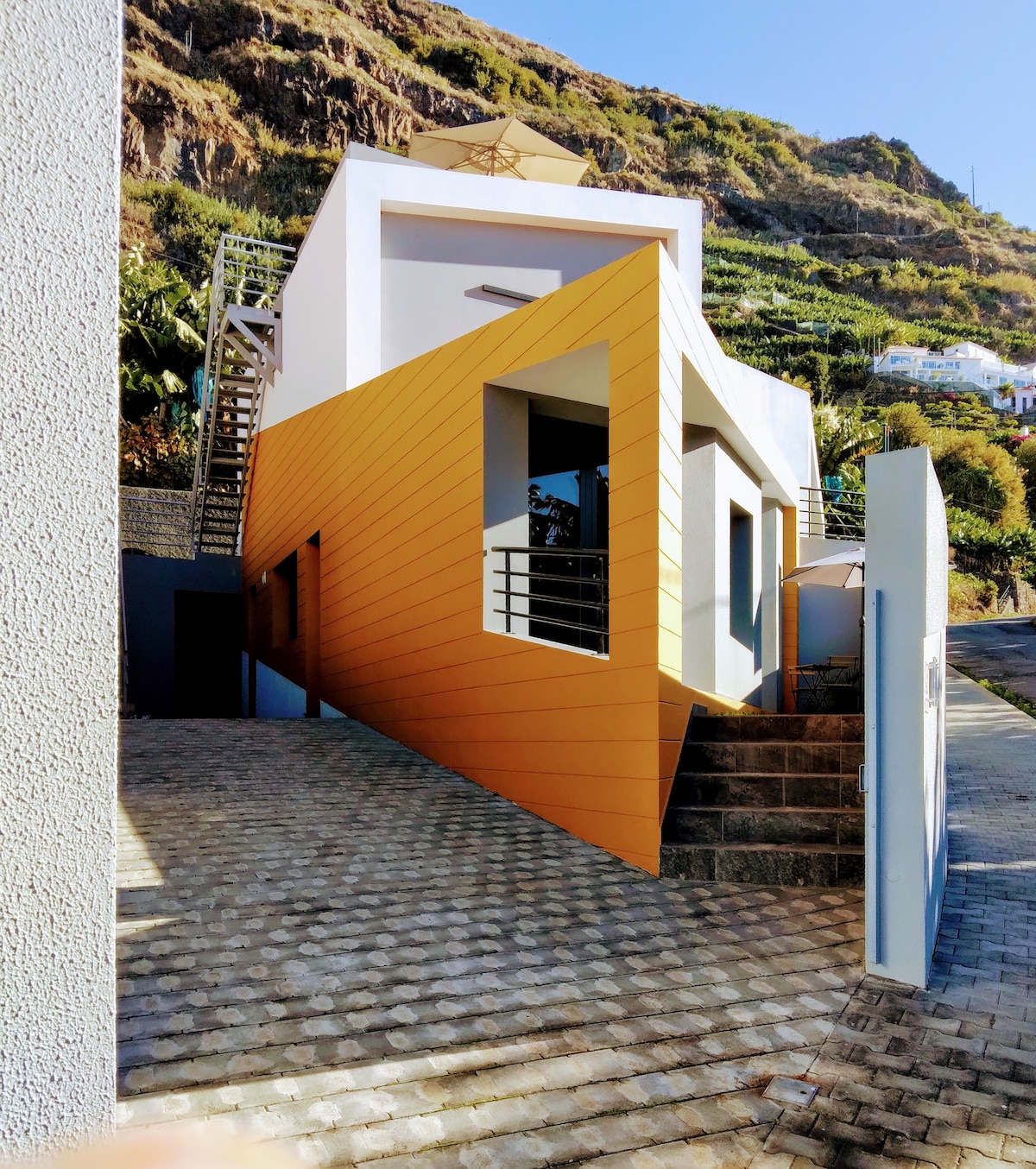 The exterior of a modern building is showcased, featuring a striking orange facade that contrasts with the surrounding natural landscape. The structure is designed with clean lines and large windows, complemented by a staircase leading to an elevated entrance.