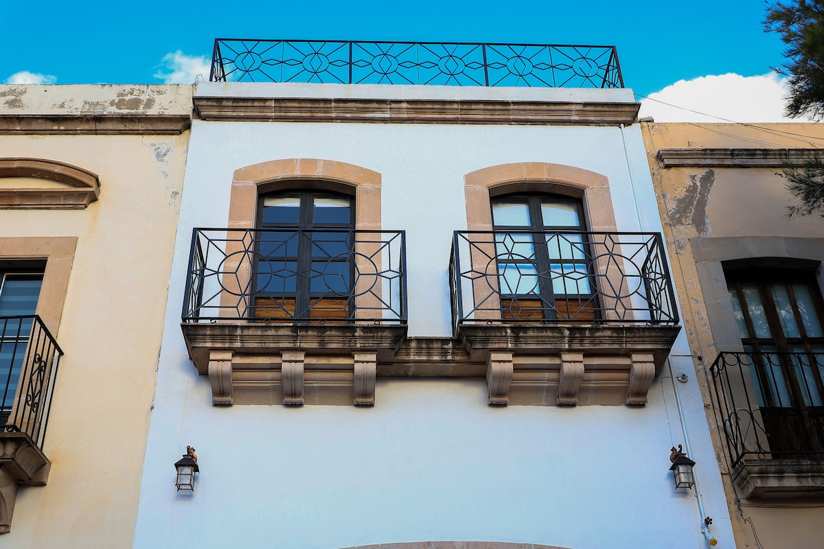 A classic building façade reveals two balconies adorned with decorative iron railings. Above, a roof terrace offers an unobstructed view of the sky. The structure is painted in soft white, contrasting with the intricate details of the neighboring buildings.