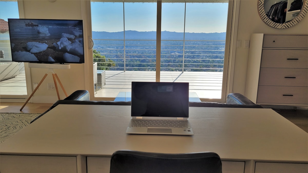 An office area is positioned in front of a large glass door that offers sweeping views of the mountains. A laptop sits on a white desk, while a television screen displays a landscape in the background. Natural light floods the space, highlighting a minimalist design.
