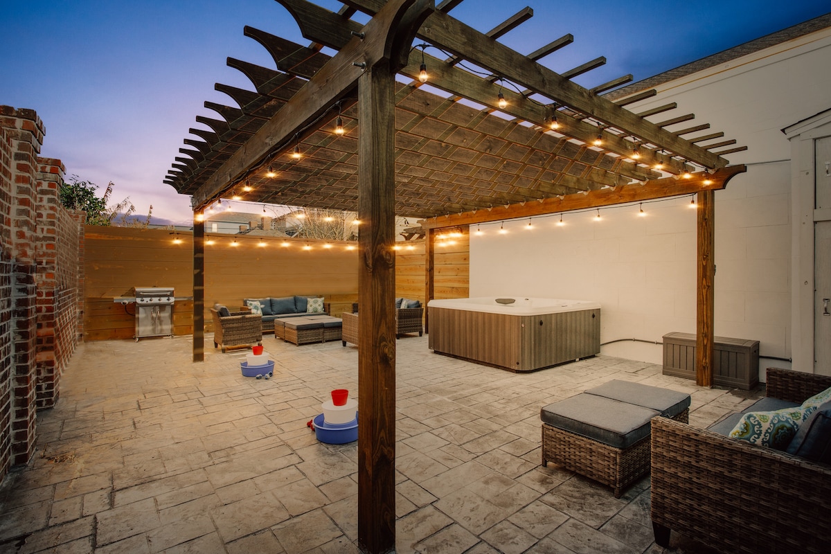 An outdoor patio area is enclosed by wooden fencing, featuring a wooden pergola adorned with string lights. Comfortable seating arrangements are placed around an eight-person hot tub, with a grill set up on one side. Two bowls are positioned on the ground.