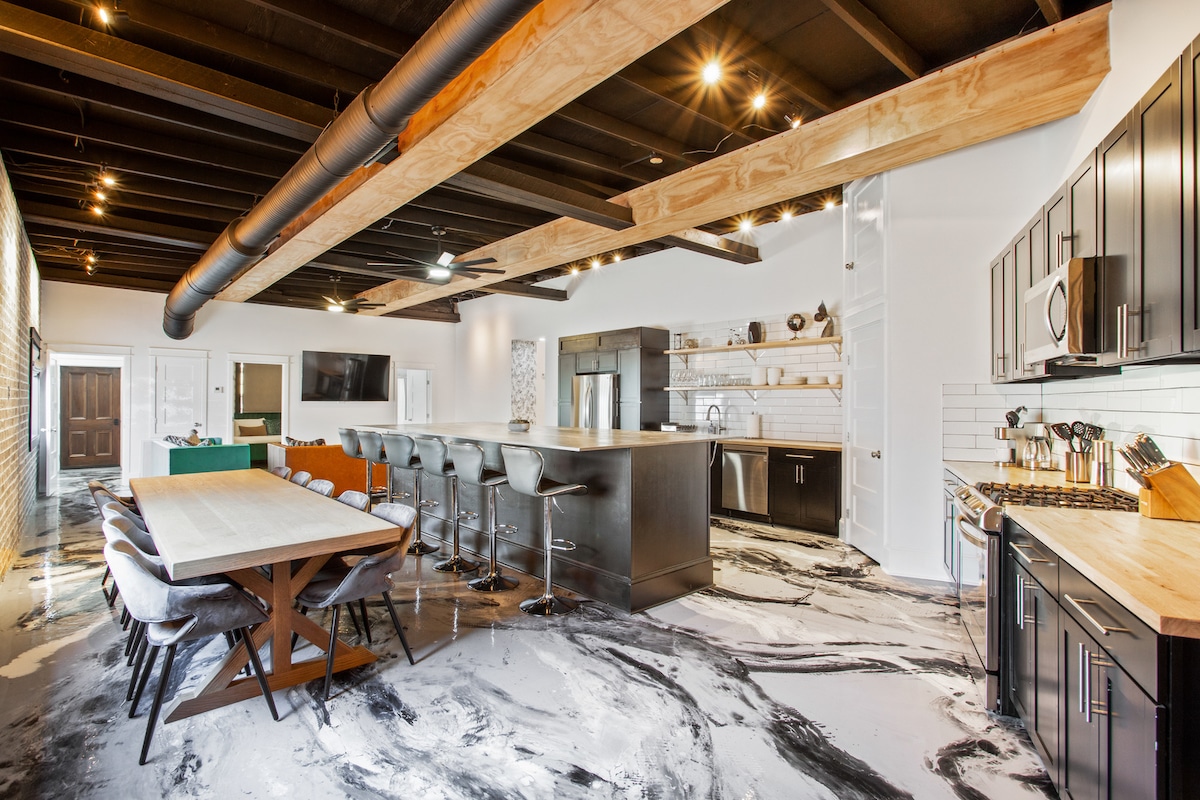 An open-concept kitchen and dining area is showcased with a spacious dining table set for six. The kitchen features sleek black cabinetry, high-end appliances, and a large island with seating for four. A modern design is highlighted by the dark ceiling beams and polished concrete flooring.