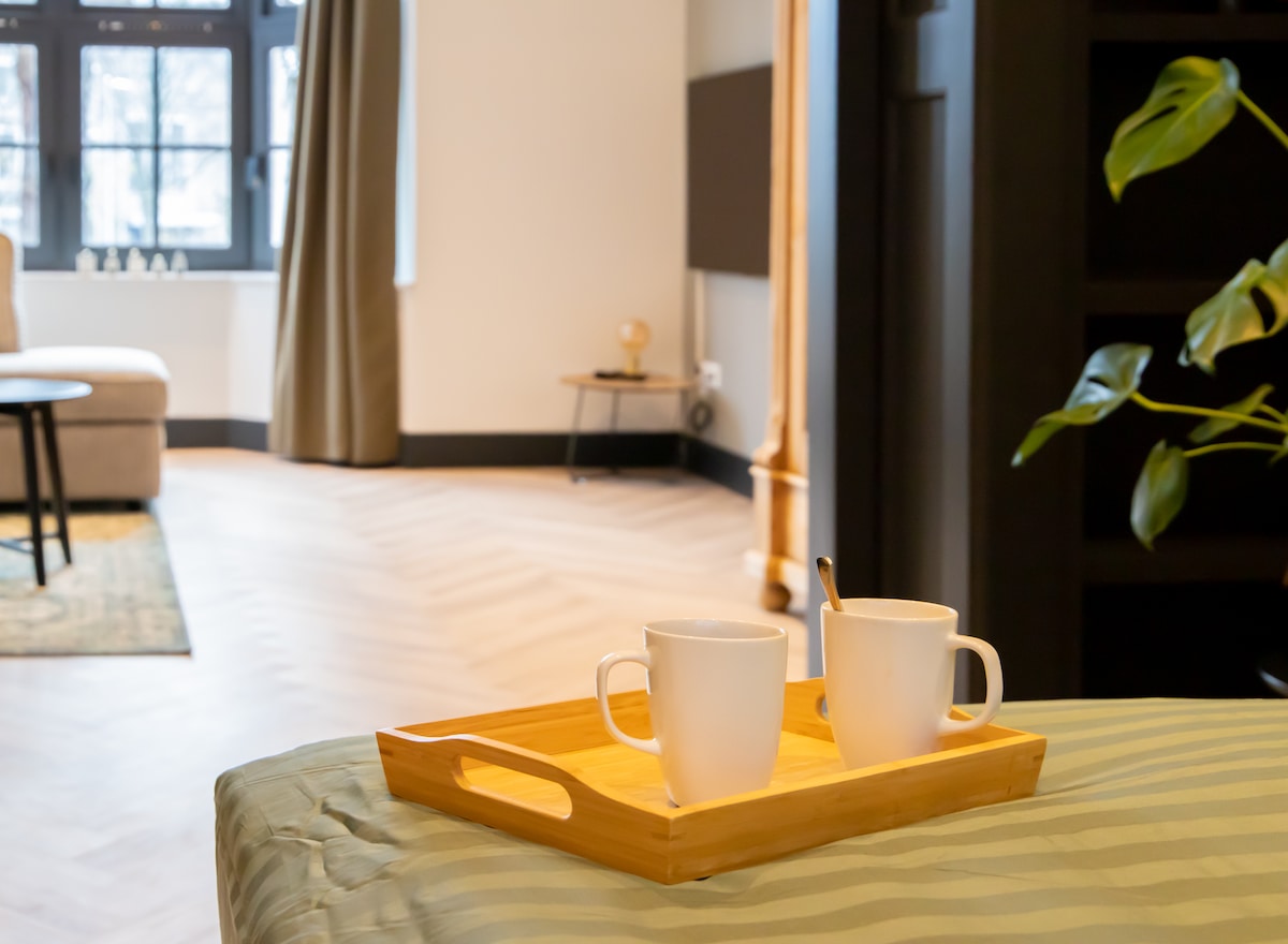 A wooden tray holds two white cups, positioned on a soft bed with a patterned cover. In the background, the spacious living area is visible, featuring large windows, curtains, and a cozy seating arrangement.