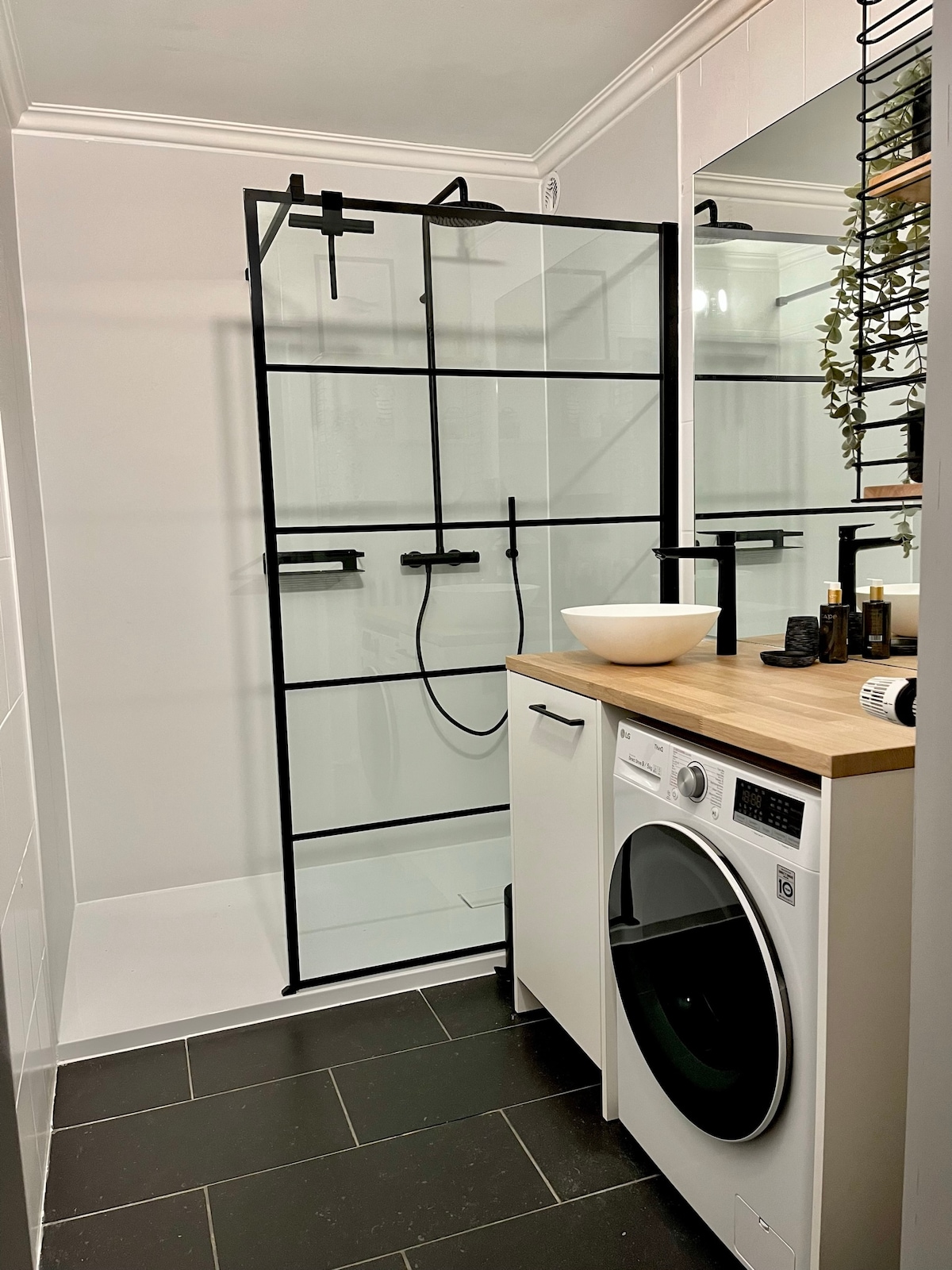 A modern bathroom features a sleek black-framed glass shower enclosure with a minimalist design. Adjacent, a compact laundry unit with a washing machine and wooden countertop provides functional space. The room is illuminated by soft lighting, enhancing the neutral color palette of the tiles.
