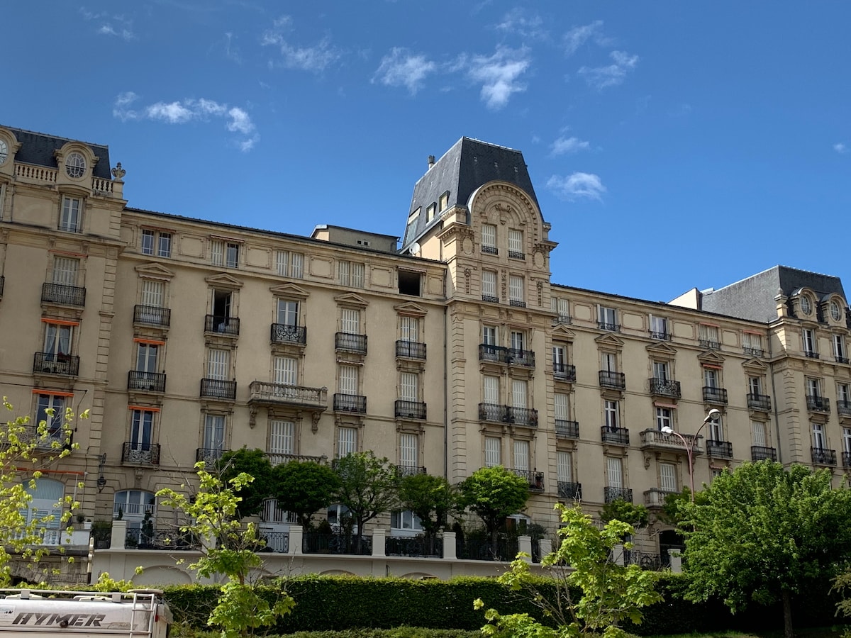 The exterior of a charming residential building is displayed, featuring a combination of balconies and large windows. A mix of architectural styles is evident, with a prominent rooftop and green foliage surrounding the base, enhancing the tranquil setting.