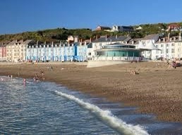 A coastal view features a beach with gentle waves lapping at the shore. Colorful buildings line the waterfront, with a modern structure in the foreground. The scene is framed by a green hillside under a clear blue sky, inviting visitors to enjoy the seaside atmosphere.