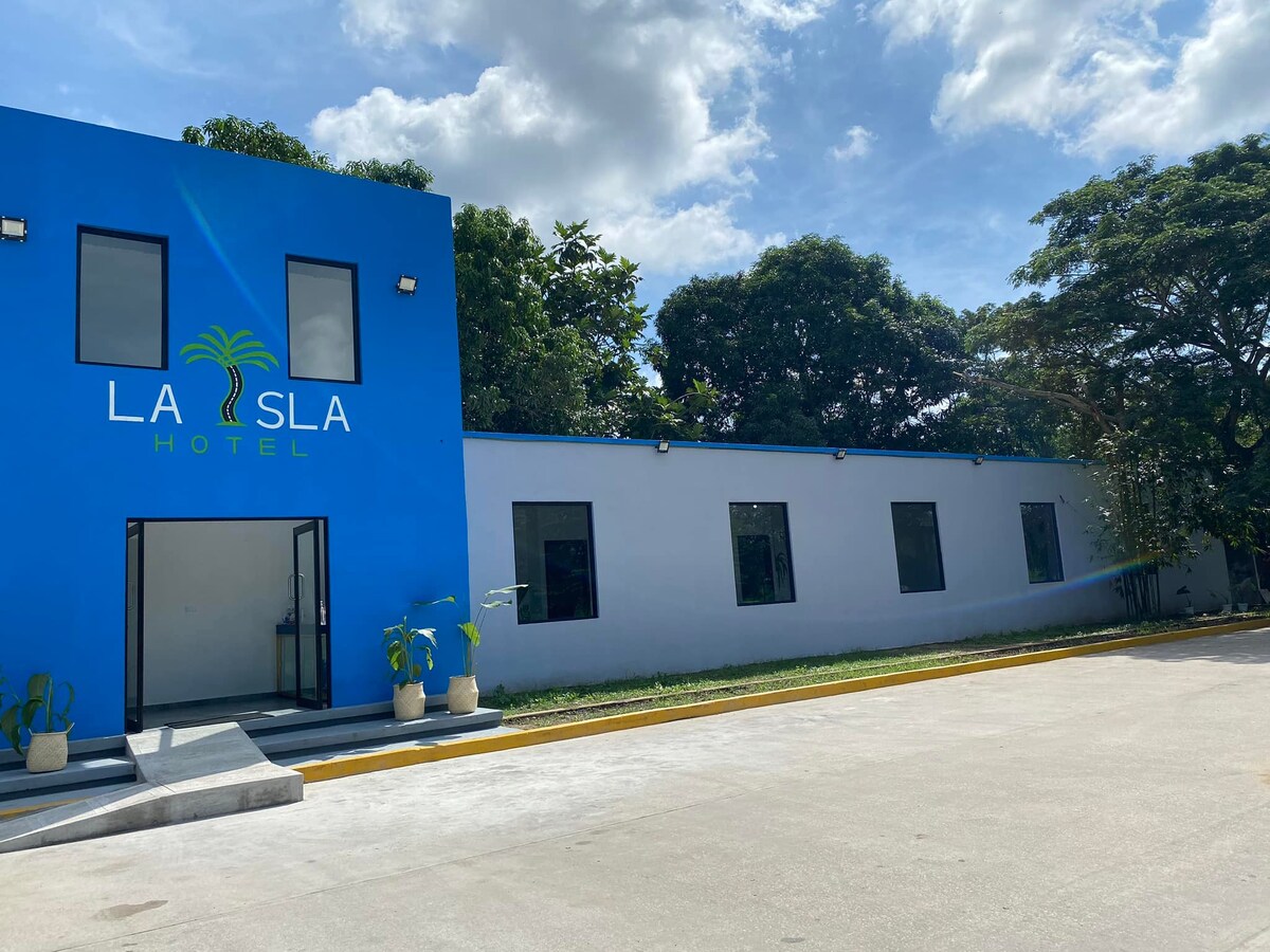The exterior of a modern hotel is featured, showcasing a vibrant blue facade with green signage. Large windows are visible along one side of the building, framed by lush greenery, and the entrance is marked by a simple, welcoming path and potted plants.