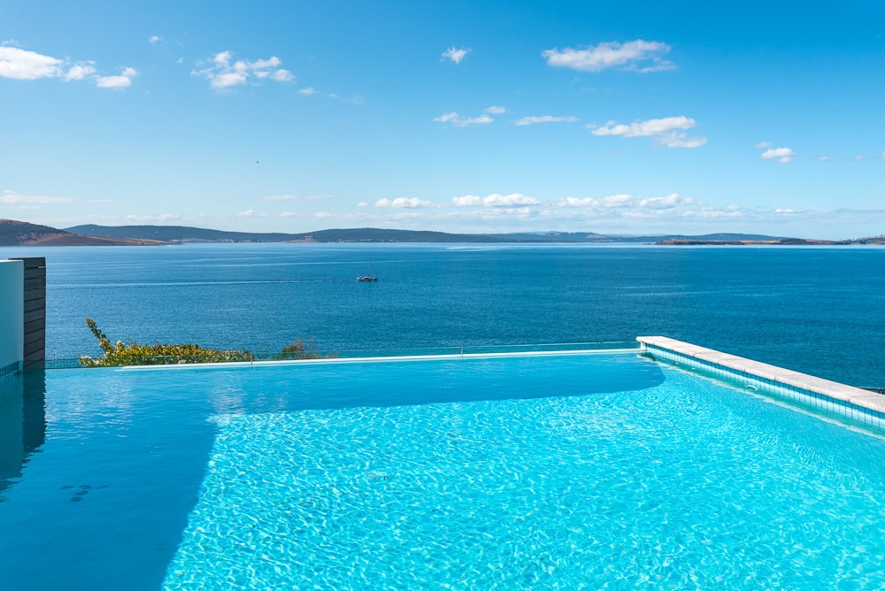 An infinity pool appears to seamlessly blend with the sparkling waters of the Derwent River, providing a refreshing escape. The expansive views of the horizon and gentle waves create a serene atmosphere under a clear blue sky.