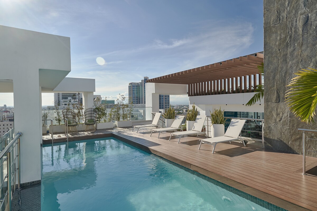 An infinity pool is visible, surrounded by a sun deck featuring several lounge chairs. Lush greenery and city views are showcased in the background, with clear blue skies enhancing the serene atmosphere of the rooftop space.