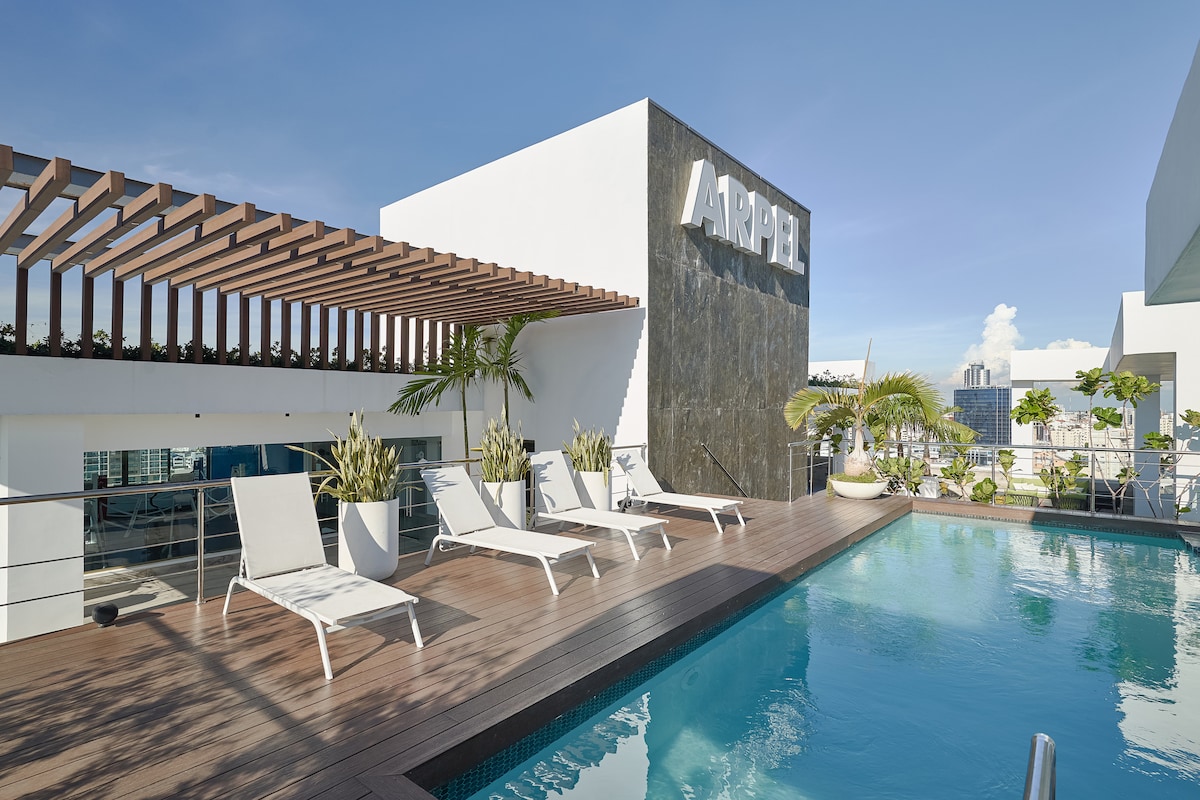 An outdoor area showcases an infinity pool surrounded by sun loungers on a wooden deck. Lush plants and a modern wall with the name 'ARAPEL' are visible in the background, under a clear blue sky, creating an inviting atmosphere.