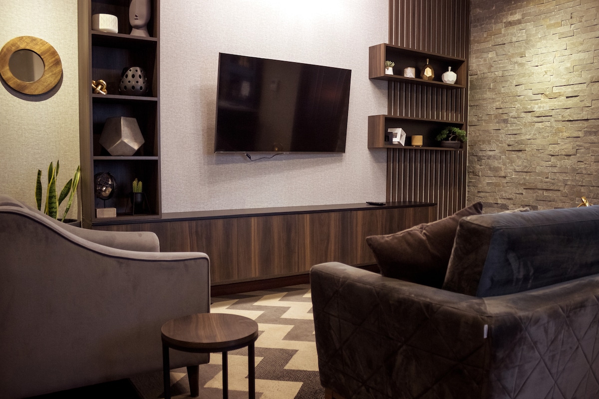 A modern living area is highlighted by a large flat-screen TV mounted on a textured wall. Two comfortable sofas face the TV, with a small round table positioned between them. Decorative shelves and plants add subtle touches of style to the room.