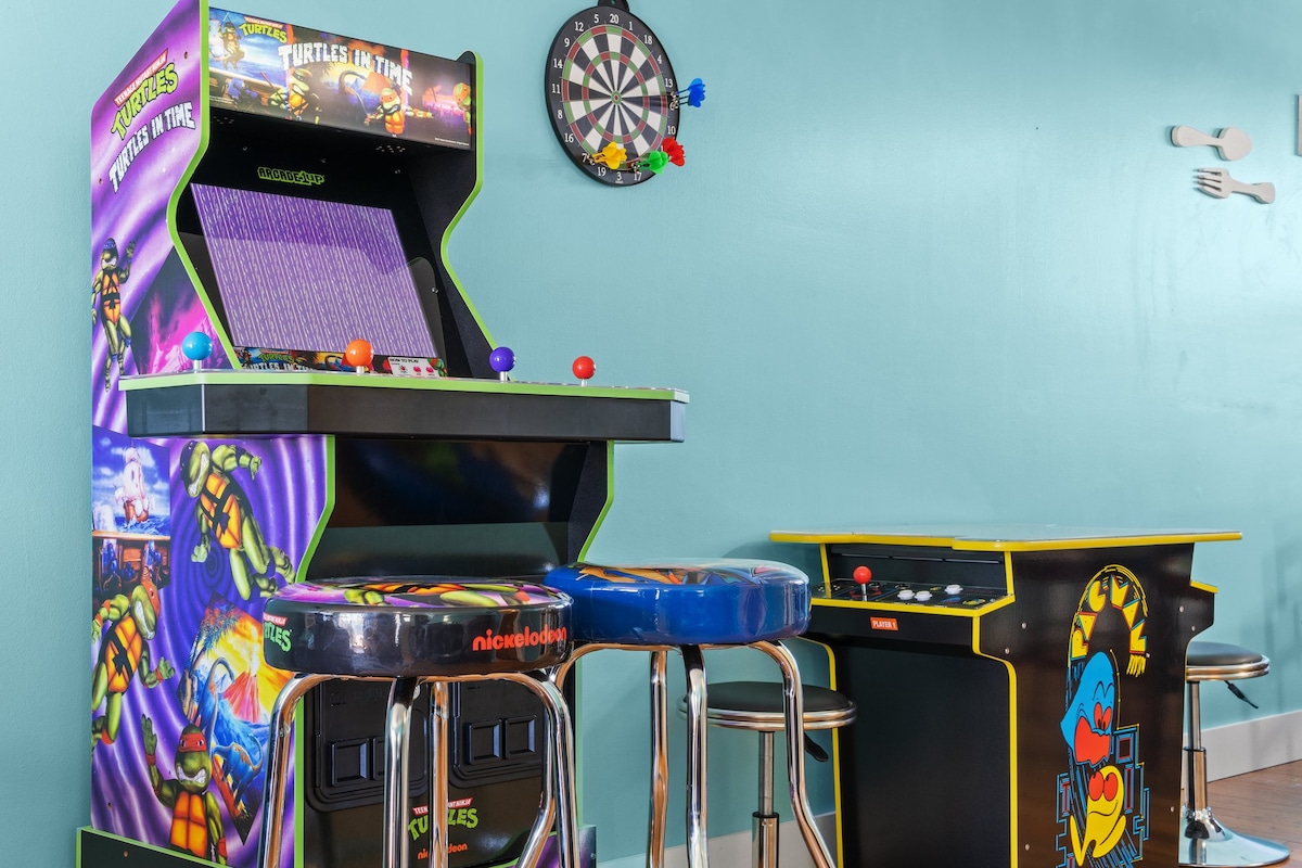 An arcade game featuring Teenage Mutant Ninja Turtles is positioned against a teal wall, accompanied by colorful bar stools. A magnetic dartboard is mounted nearby, providing additional entertainment options.