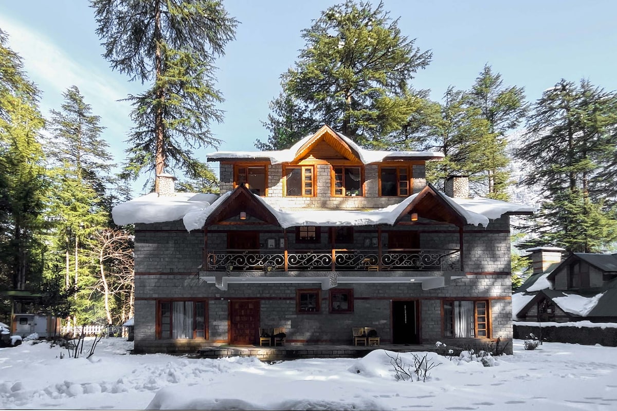 A charming gray wooden structure is framed by snow-covered trees. A prominent balcony with wooden railing graces the upper level, while multiple windows invite natural light. The ground floor features a combination of doors and windows, enhancing the cozy, lodge-like ambiance of the home.