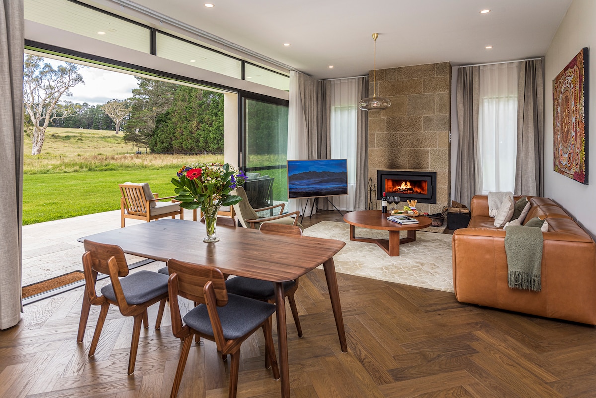 An open living area is shown with large glass doors leading to a picturesque outdoor setting. A leather couch faces a coffee table, adjacent to a modern fireplace. A dining table with four chairs stands nearby, and an entertainment setup includes a flat screen TV.