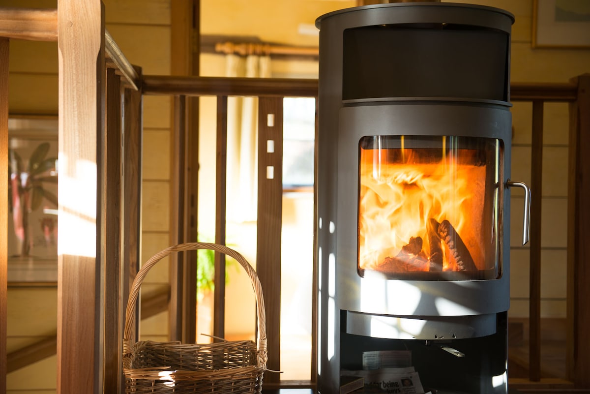 A modern wood-burning stove stands prominently in the room, flames flickering warmly within its glass enclosure. A woven basket for firewood is positioned beside it, blending with the natural wooden elements of the interior.
