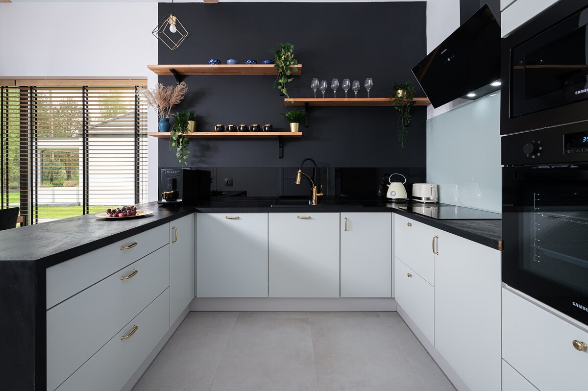 A modern kitchen is presented, featuring light grey cabinetry paired with dark countertops. Open shelving displays decorative elements alongside glassware. A microwave and toaster are positioned for easy access, while large windows allow natural light to fill the space, creating a bright and functional environment.