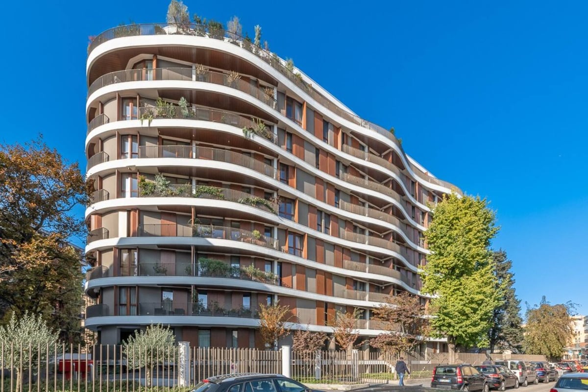A modern apartment building is depicted with a curved design, featuring multiple balconies adorned with greenery. The structure showcases a blend of materials and colors, surrounded by trees and parked cars, set against a clear blue sky.