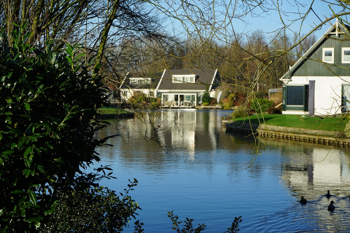 A tranquil view of a villa reflected in the calm waters of the river, framed by greenery on the bank. The surrounding landscape features gently sloping vegetation, enhancing the serene atmosphere of the location.