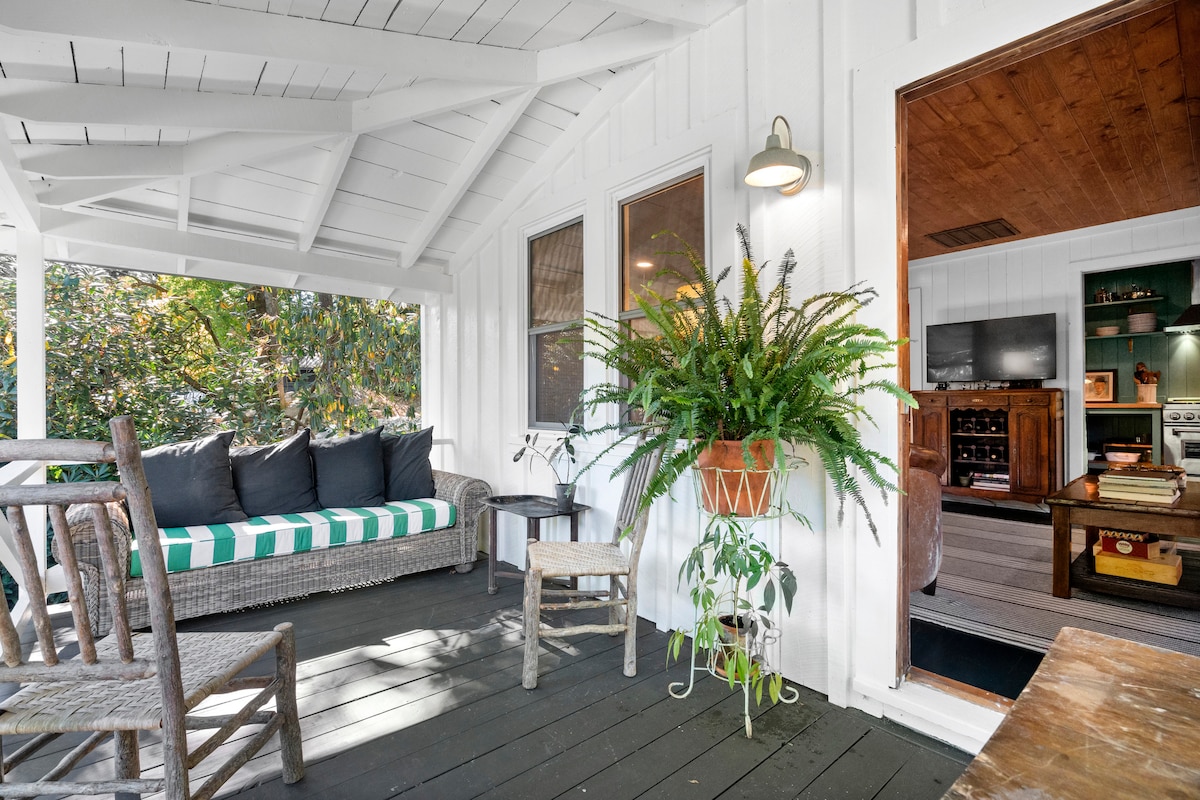 A screened front porch features a comfortable seating area with a spacious couch adorned in green and white stripes. A small table accompanies the couch, while large windows allow natural light to fill the space. Lush greenery is present in a hanging planter.