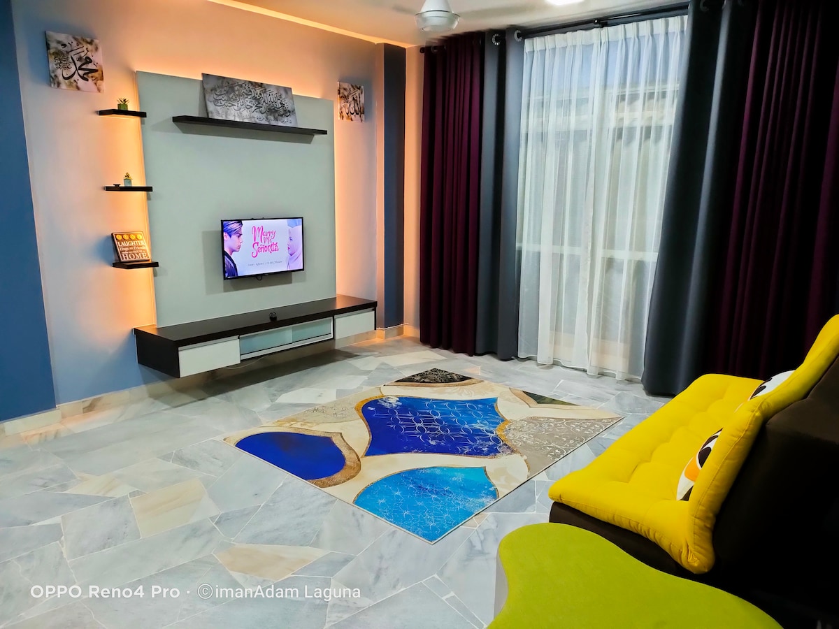 A living area features a wall-mounted TV on a sleek cabinet beneath decorative shelves. Large windows with floor-to-ceiling curtains allow natural light. A colorful area rug adds a touch of design, while a yellow sofa is positioned alongside a dark-colored sectional sofa.