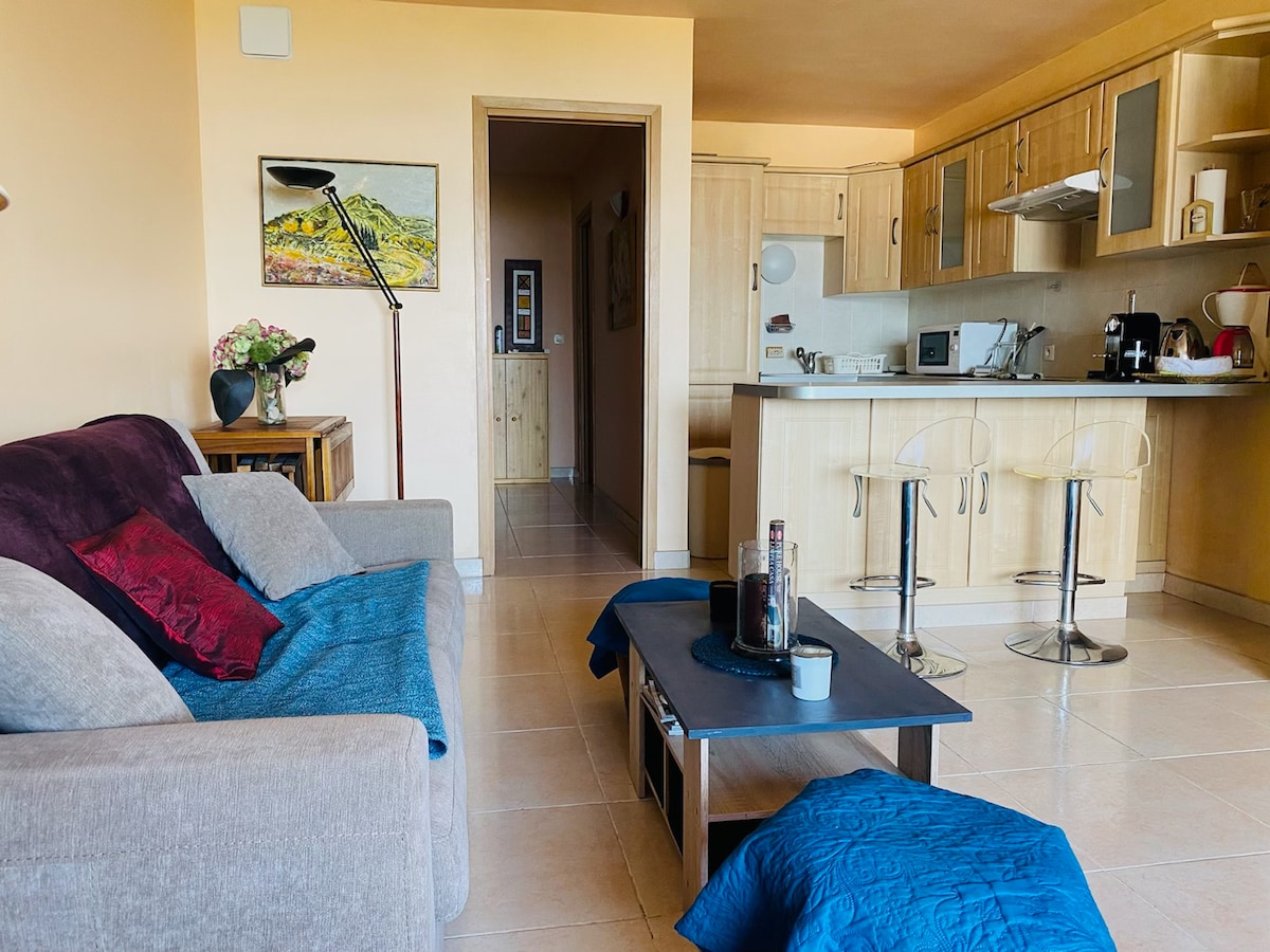 A spacious living area is visible, featuring a soft gray sofa adorned with colorful cushions. A small wooden table sits in front, accompanied by two stylish bar stools. The kitchen area showcases light wooden cabinetry and modern appliances, creating a welcoming space for relaxation and dining.