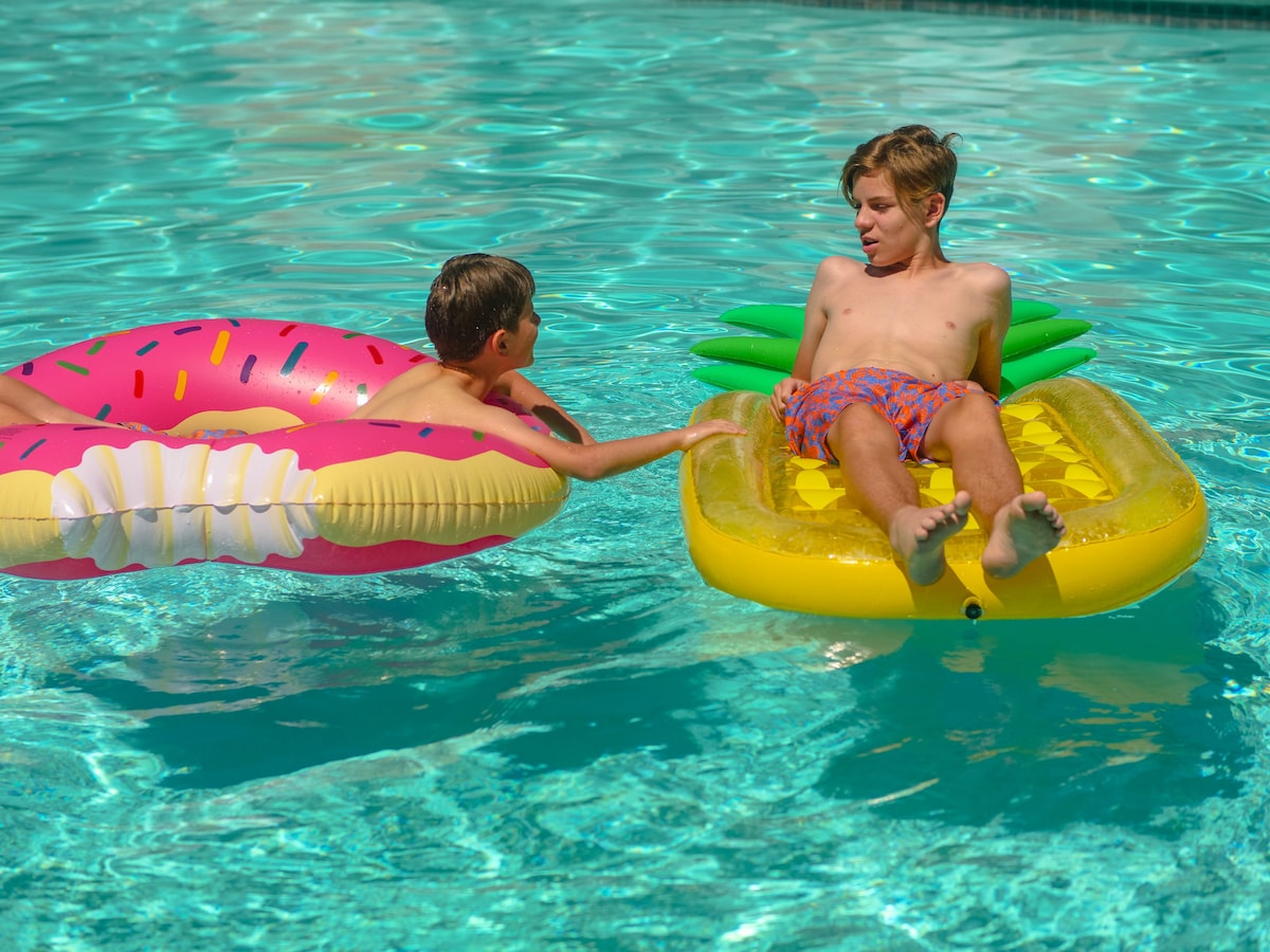 Two children are enjoying a sunny day in a pool. One child is seated on a yellow inflatable float shaped like a pineapple, while the other is on a pink donut-shaped float. The clear blue water reflects light, creating a vibrant atmosphere.