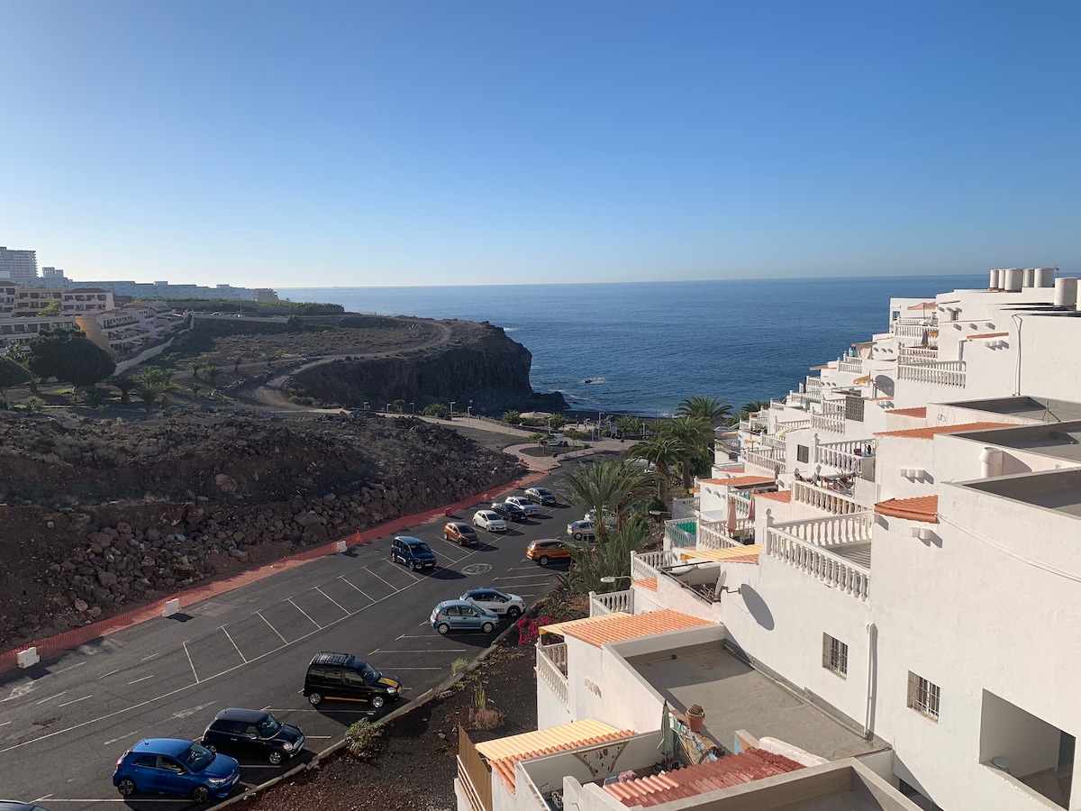 A coastal view is presented, featuring the ocean meeting a clear blue sky. Nearby, a parking area with various cars is visible, alongside a landscaped pathway leading towards the rocky shoreline. The scene captures the tranquility of the seaside environment.