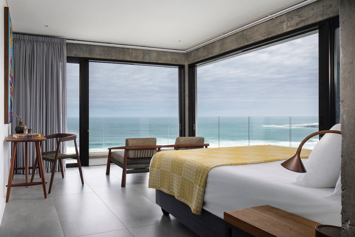 A modern bedroom features large glass windows framing expansive ocean views. A spacious bed is adorned with a textured yellow throw. Two stylish chairs and a small table are positioned nearby, while soft curtains add a touch of privacy.