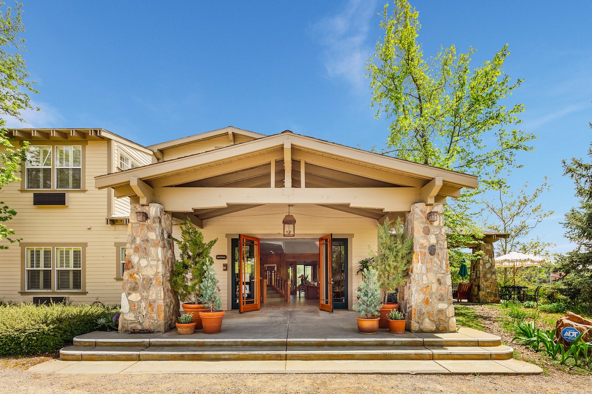 The entrance features a covered porch framed by stone columns and wooden beams. Bright orange doors open to a welcoming interior, surrounded by lush greenery and potted plants. Natural light filters through large windows, enhancing the inviting atmosphere.
