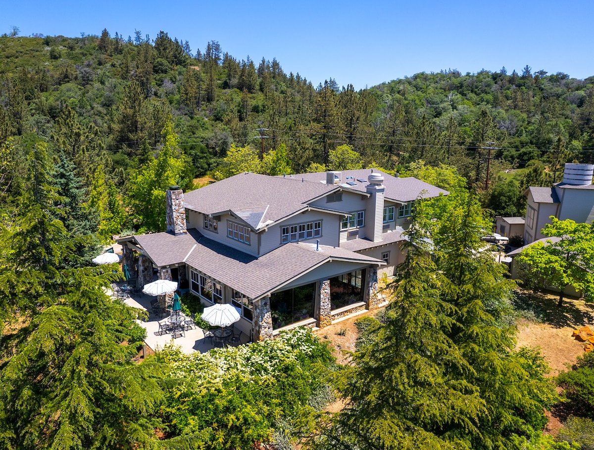 An expansive cottage complex is surrounded by lush greenery, with outdoor seating areas featuring umbrellas. The building's stone accents complement the natural landscape, creating a harmonious blend with the tranquil outdoors in a peaceful, private setting.