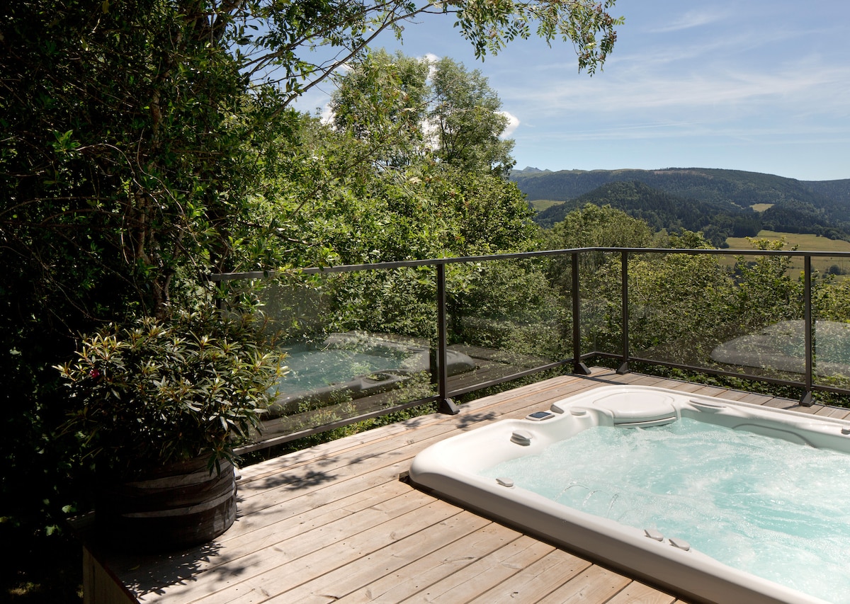 An outdoor spa is situated on a wooden deck, surrounded by greenery. A glass railing provides a clear view of the surrounding hills and valleys under a clear blue sky.