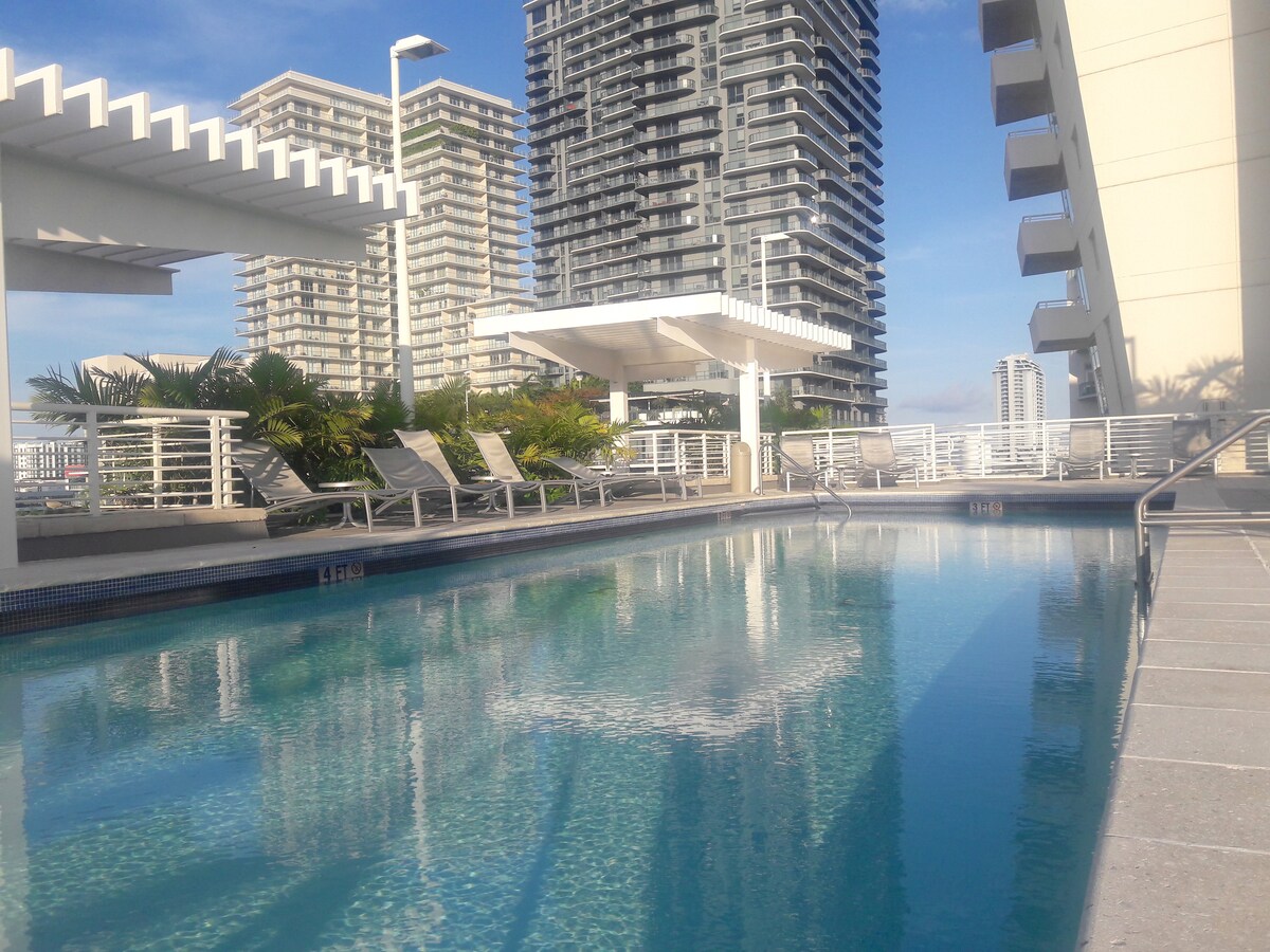 A community pool area is featured, surrounded by lounge chairs and palm trees. Modern high-rise buildings reflect in the serene water under a clear blue sky. A shaded seating area with a pergola provides a welcoming space for relaxation.