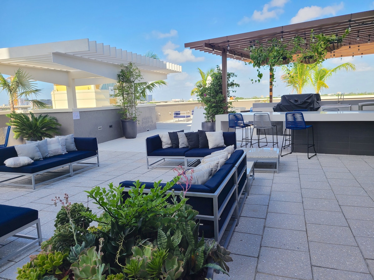 An outdoor lounge area features comfortable seating on a tiled surface, surrounded by lush greenery and palm plants. High-top seating at a bar overlooks the skyline, while shaded areas provide a relaxing space. Bright sunlight filters through the open spaces, enhancing the atmosphere.