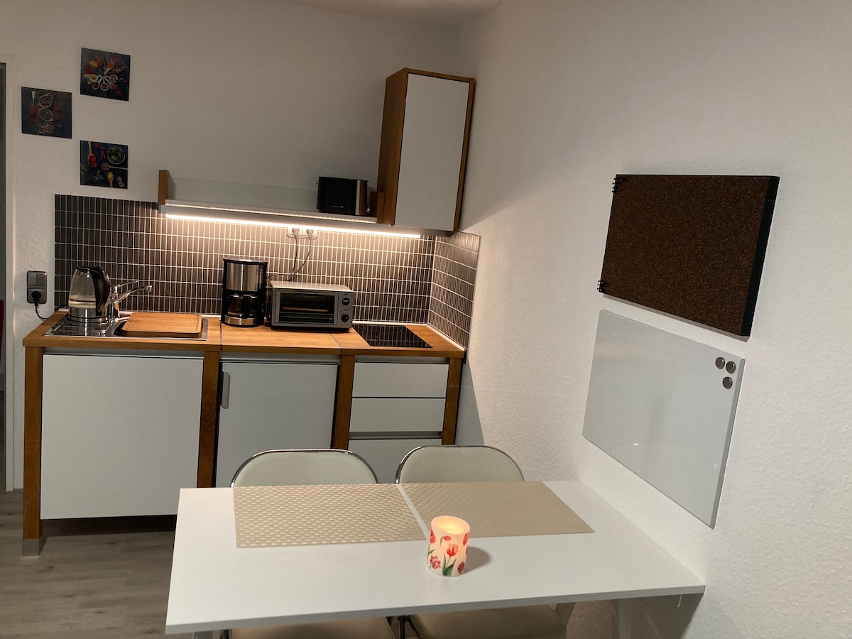 A compact kitchen area is equipped with essential appliances including a stove, mini oven, and refrigerator. Cabinets are set against a tiled backsplash. A dining table with two chairs is positioned in front, featuring placemats, while a small candle adds a touch of warmth.