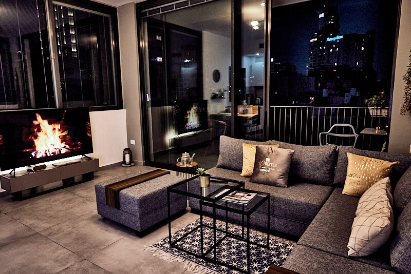 An open-concept living area is presented with a grey sectional sofa and a low coffee table. A large television sits on a modern console, while a faux fireplace offers a cozy visual element. Floor-to-ceiling glass doors showcase the city skyline at night.