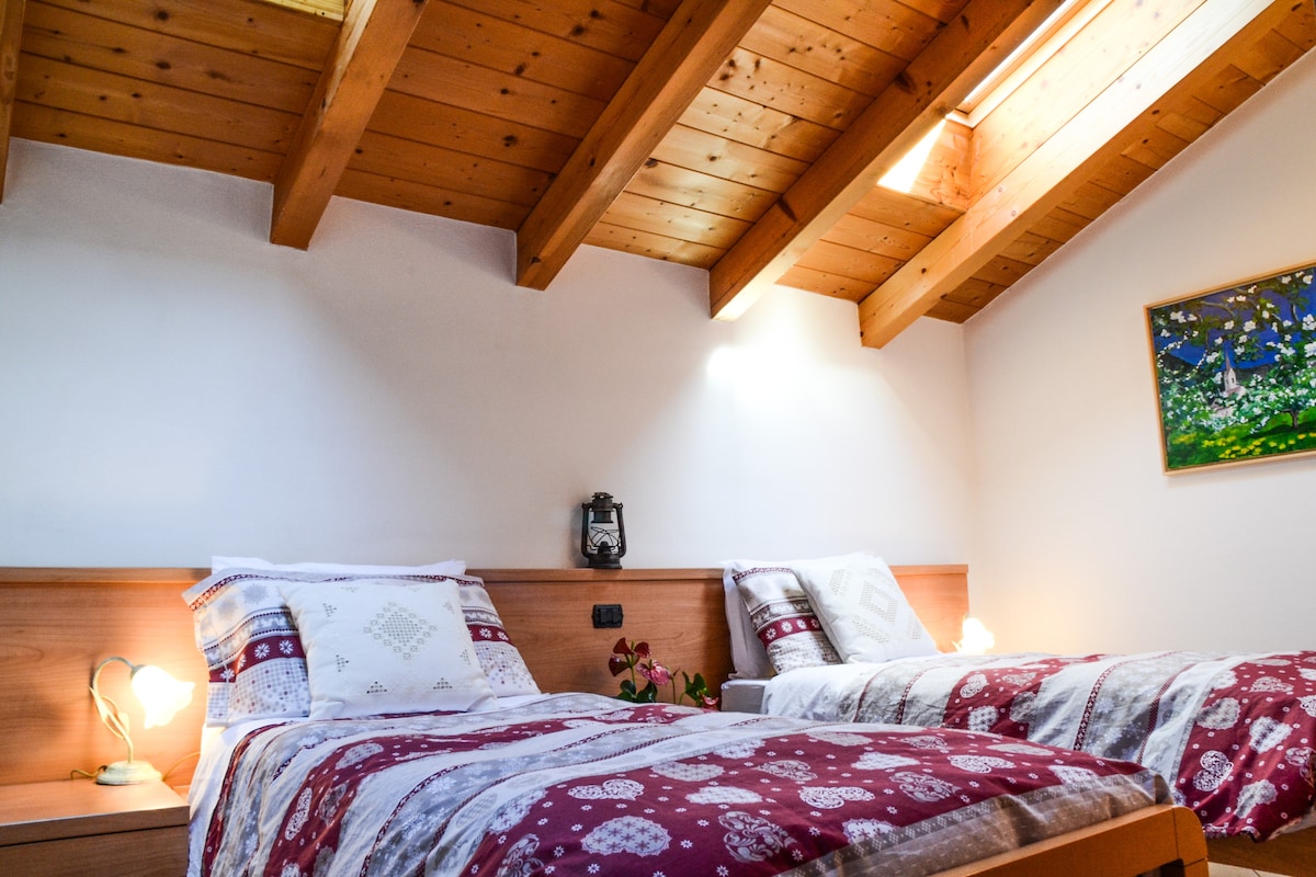 A room featuring two beds with patterned bedding is illuminated by natural light streaming through a skylight. The ceiling is adorned with wooden beams, creating a warm atmosphere. A bedside table holds a lamp, and a small decorative piece is positioned on the wall.