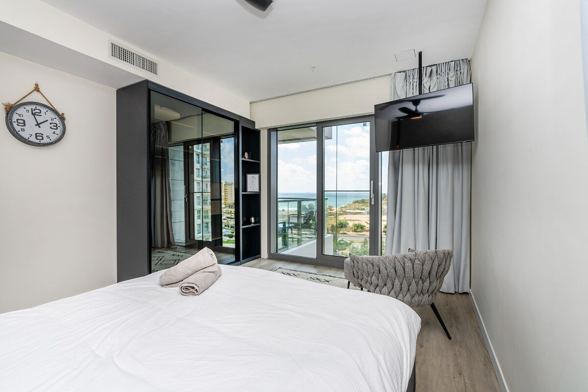 A modern bedroom is displayed, featuring a spacious bed with a white duvet and neatly rolled towels. A flat-screen TV is mounted on the wall, while large windows offer views of the outdoor landscape. A cozy armchair is positioned beside the bed.
