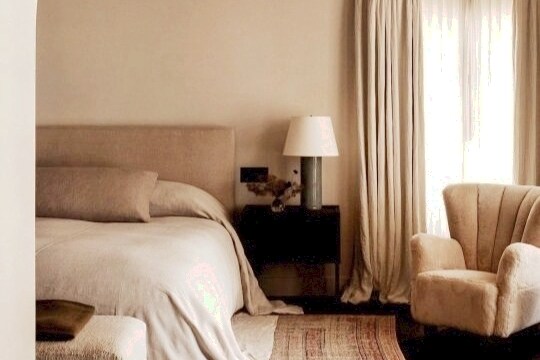 A serene bedroom is presented with a spacious bed dressed in neutral tones. A bedside lamp sits atop a dark nightstand, and a textured armchair is placed nearby. Soft curtains allow natural light to filter in, complementing the calming atmosphere.
