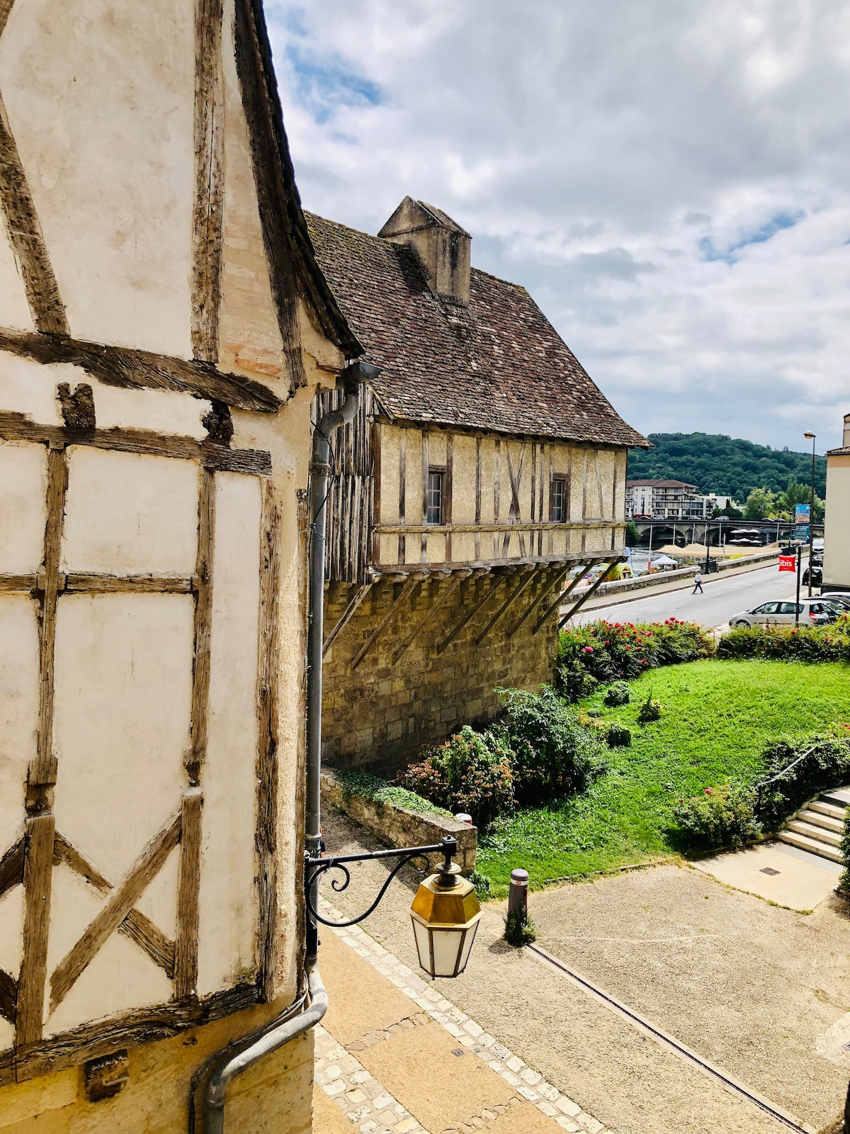 The image captures a view of half-timbered architecture alongside a lush green garden. A historic building extends over the ground, supported by wooden stilts. The scene is framed by gentle clouds and smooth roadways, hinting at a vibrant neighborhood setting.