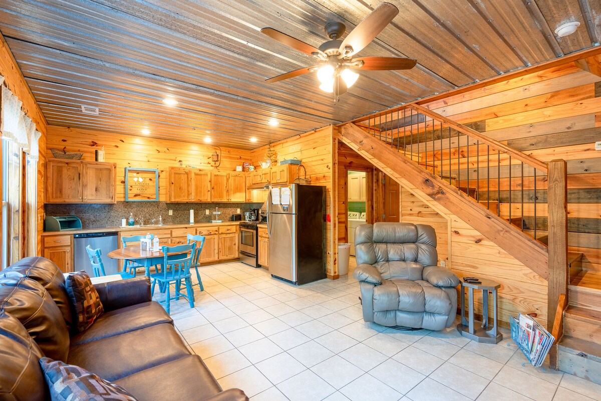 An open living area features a cozy seating arrangement with a brown sofa and a reclining chair. A dining table with blue chairs is positioned nearby, alongside a well-equipped kitchen with wooden cabinetry and modern appliances. Natural light fills the space, highlighting the wood-paneled walls.