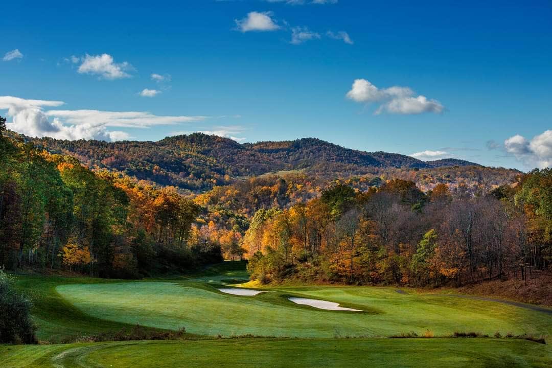 A scenic view captures a golf course surrounded by rolling hills and vibrant autumn foliage. The landscape features well-maintained green grass with sand traps visible in the foreground, under a clear blue sky dotted with clouds.